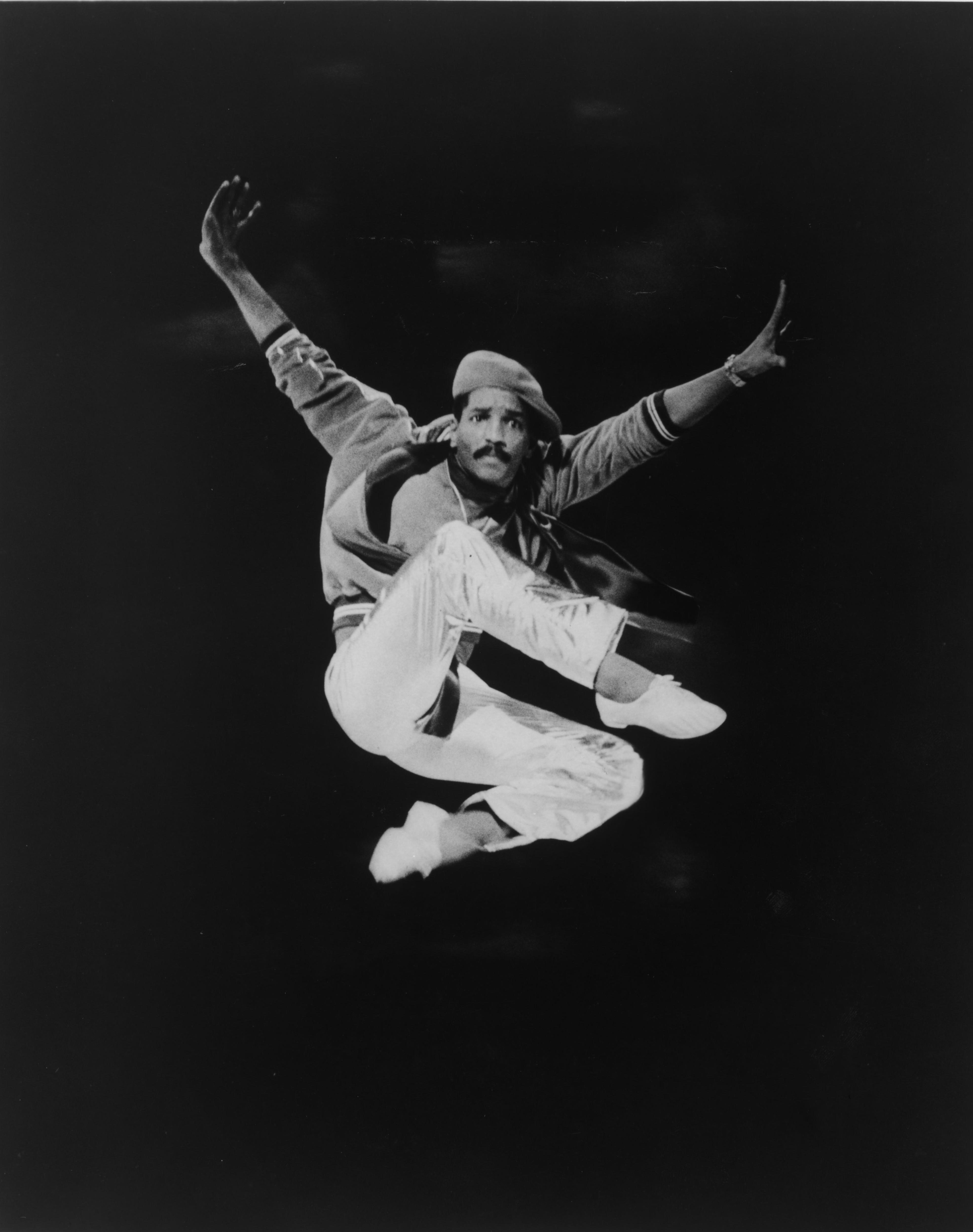 A man is jumping in the air with his arms in a V. His legs are crossed. It's black & white. He's wearing a pageboy hat and has a mustache.