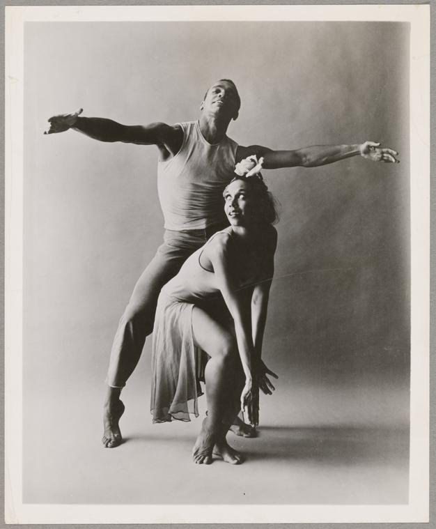 A man poses with a woman in this black and white photo. The man's arms are outstretched and the woman is squatting in front of him with her arms on her knees.