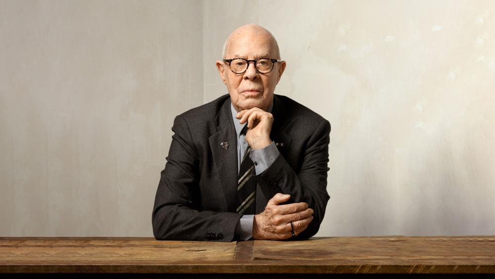 A man in a suit sits at a wood table. His hand is on his chin.