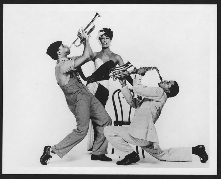 Black and white photo of Carl Bailey, Debora Chase, and Gary DeLoatch in Alvin Ailey's "For 'Bird' - With Love." Bailey and DeLoatch are posed playing trumpets, while Chase stands in the center wearing a glamorous outfit with a feathered headpiece. Photo by Jack Mitchell, 1991.