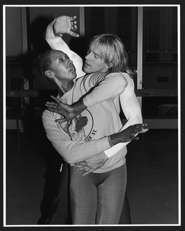 Judith Jamison and Alexander Godunov are captured in an intense and focused rehearsal for Alvin Ailey's "Spell" in 1981. Jamison is wearing a sweatshirt with an image on the front and tights, while Godunov is behind her, both engaged in a complex and expressive dance pose. The photograph conveys the dedication and artistry involved in their performance, with both dancers deeply immersed in their movements. The background suggests a studio setting, adding to the atmosphere of preparation and practice.