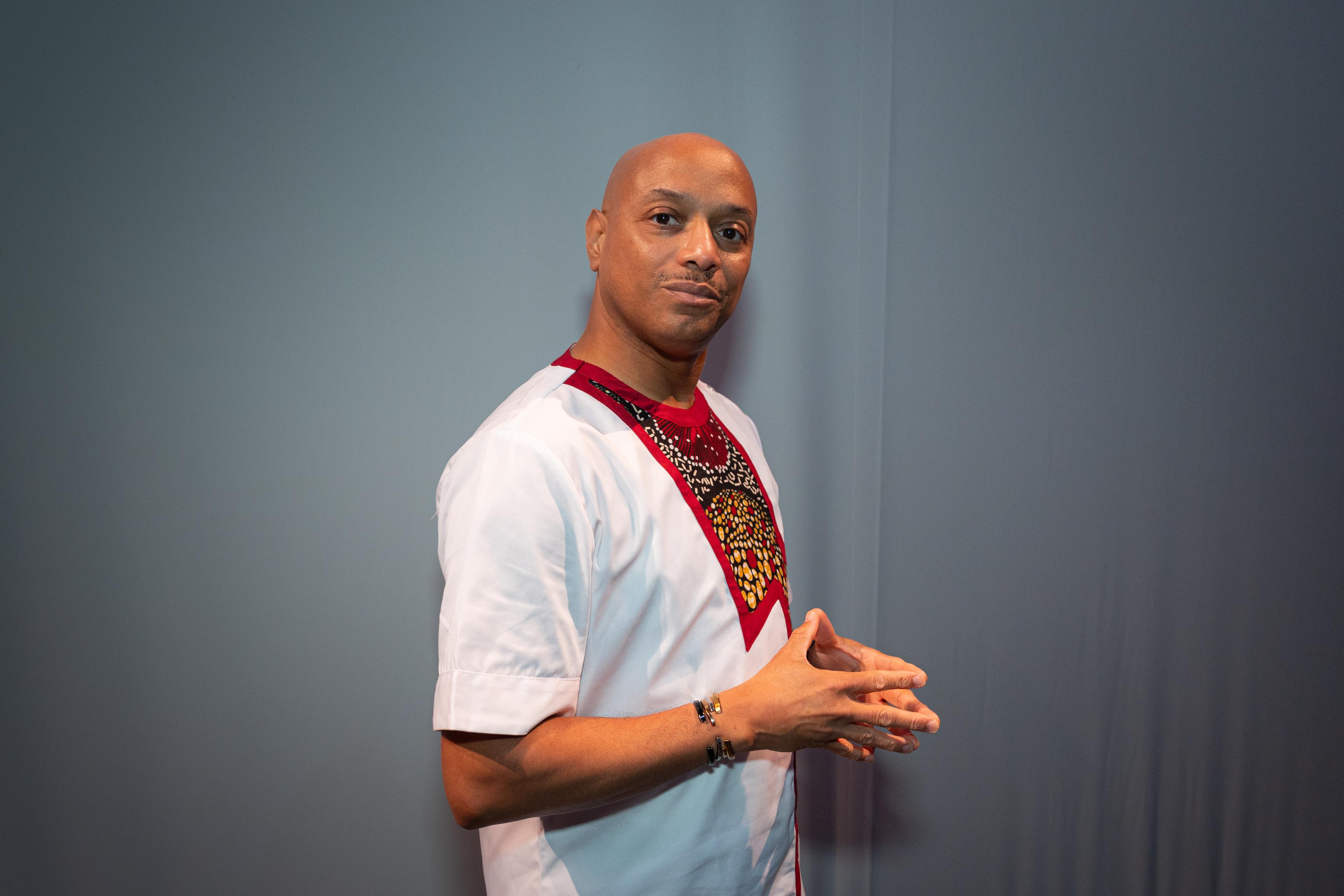 A black man with no hair, stands sideways with hands clasped in front of him.