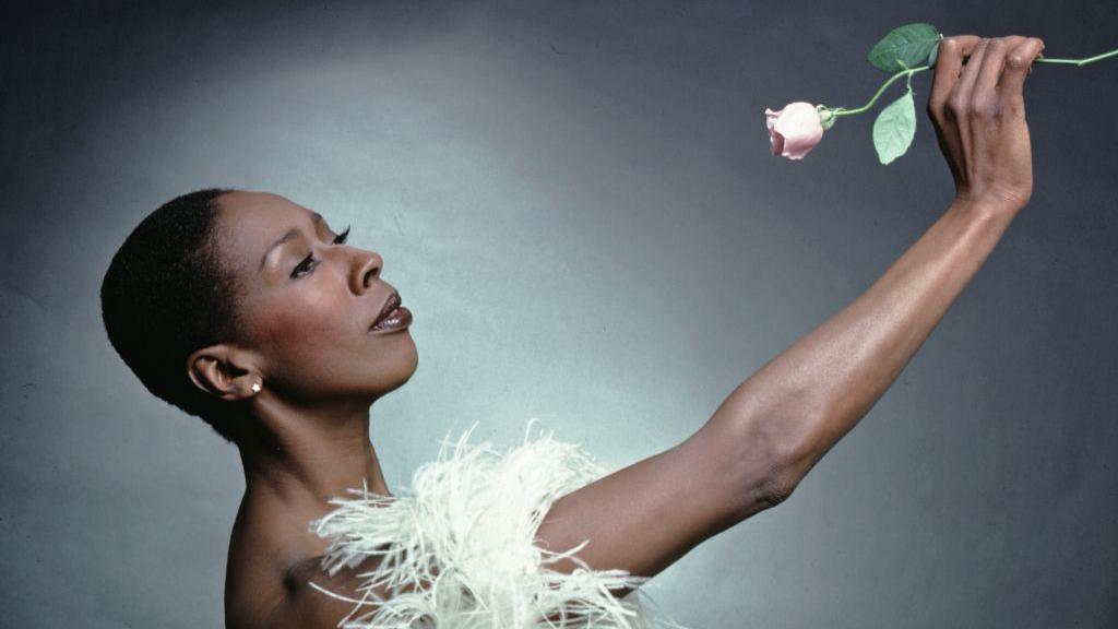 Judith Jamison, a black woman, holding a pink rose and wearing white feathers